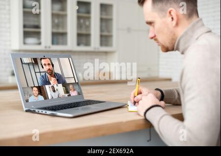 Attentive young hipster man sitting at table with laptop, working remotely from home and taking notes to notebook while on team meeting online with coworkers, looking at screen with people profiles Stock Photo