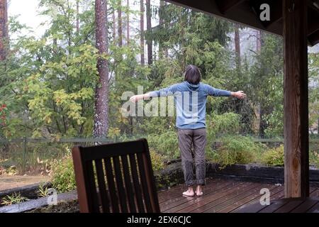 Woman spread her arms to the sides and enjoys the warm rain, standing on the veranda and looking at the forest. Unity with nature. Stock Photo