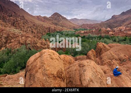 Morocco, Dades Valley near the 'Monkey Paws' rock formations, with Berber male dressed in blue. A barren landscape with lush vegetation along a river. Stock Photo