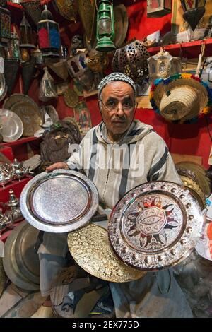 A silver smith working on his wares in a shop, showing silver plates, tea pots and trays