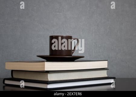 Brown cup of coffee with saucer on top of pile of books to read placed off center of frame on a clean gray background. Empty space for text Stock Photo