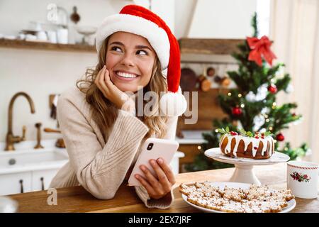 Happy nice woman in santa claus hat smiling while using mobile phone in cozy kitchen Stock Photo