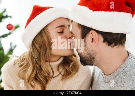 Beautiful happy couple in santa claus hats smiling and kissing indoors Stock Photo