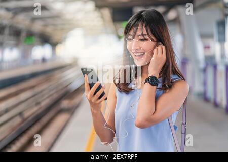Asian woman using face recognition via smart mobile phone and listening the music at Railroad Station Platform, Biometric Verification and artificial Stock Photo