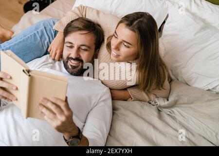 Happy young couple reading book while laying in bed at home Stock Photo