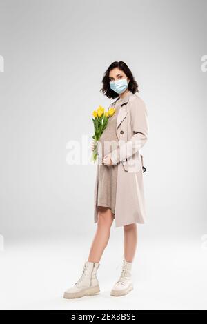 Pregnant woman in medical mask holding flowers on grey background Stock Photo