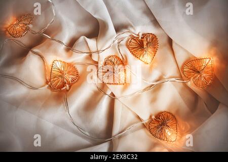 A glowing garland with heart-shaped bulbs glows with a warm light and lies on a white soft fabric, creating comfort and romance. Valentine's day. Wedd Stock Photo