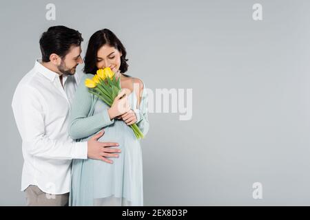 Happy man embracing pregnant wife smelling flowers isolated on grey Stock Photo