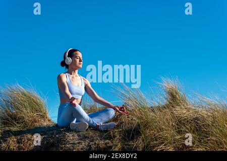Woman meditating and listening to music outside in the sun. Wearing headphones.