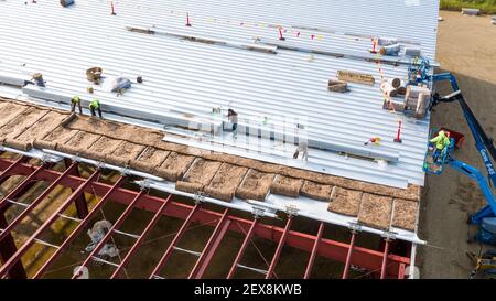 Construction workers installing insulation and roof panels on a large warehouse building. Stock Photo