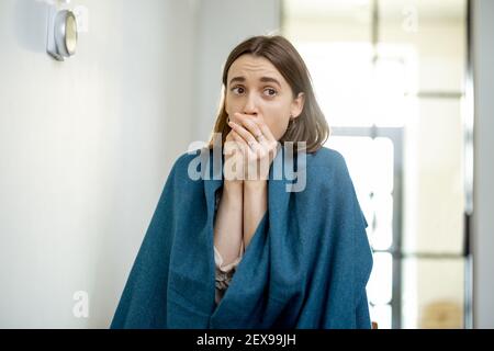 Pretty woman is cold covered with a plaid while blowing in her hands and standing near the heat regulator on the wall waiting for the temperature to rise. Smart home technology.  Stock Photo