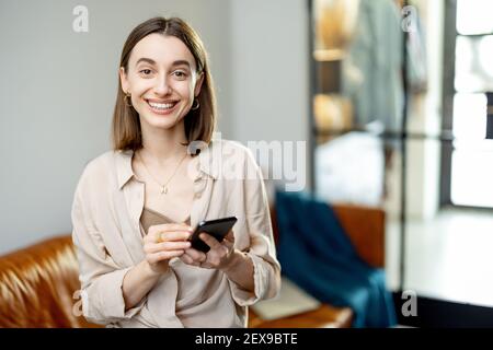 Portrait of cheerful woman using Smartphone on home interior background. Lifestyle concept. Look and smile at the camera. Stock Photo