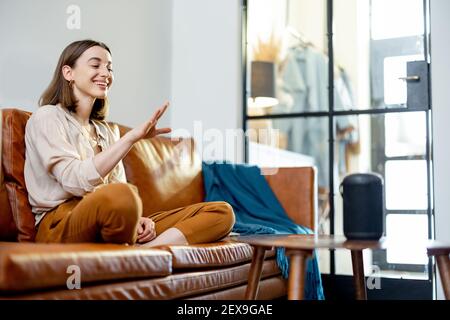 Happy pretty woman speaking with black audio assistant column on the coffee table while sitting on brown leather sofa in stylish living room. Smart home concept. Stock Photo