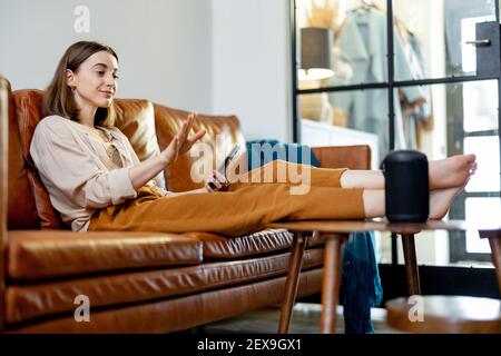 Happy pretty woman speaking with black audio assistant column on the coffee table while sitting on brown leather sofa in stylish living room. Smart home concept. Stock Photo