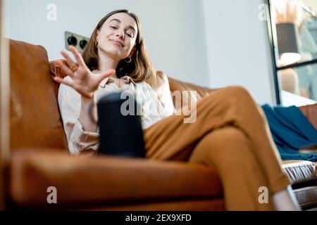 Happy pretty woman speaking with black audio assistant column on the coffee table while sitting on brown leather sofa in stylish living room. Smart home concept. Stock Photo