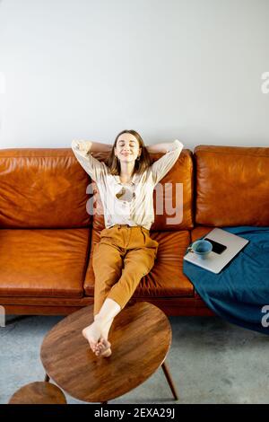 Pretty woman relaxing and dreaming on the brown sofa after working on laptop. Lifestyle and work from home concept.  Stock Photo