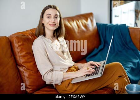 Pretty woman with laptop sitting on the brown sofa at home. Lifestyle and work from home concept.  Stock Photo