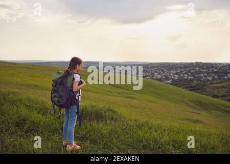 Girl tourist hiker with backpacks looks at the sunset standing on a hill in the summer. Little kid girl on foot travel adventure in nature Stock Photo