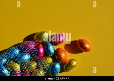 Foil wrapped chocolate Easter eggs spilling out of blue plastic net bag, isolated on bright yellow background with copy space Stock Photo