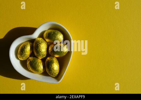 Gold foiled wrapped chocolate Easter eggs in a white heart shaped ceramic dish isolated on yellow background Stock Photo