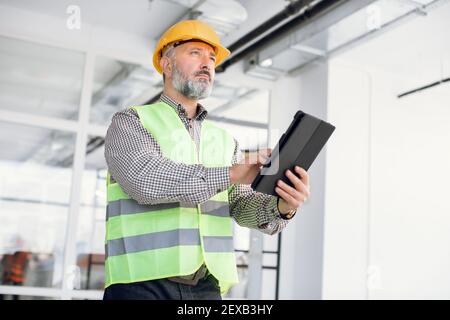 Mature inspector in hardhat working at tablet on object Stock Photo