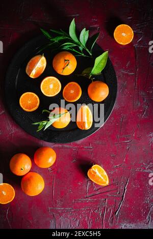 Fresh whole, halved, and quartered oranges laid out on a deep red surface and black serving platter. Stock Photo