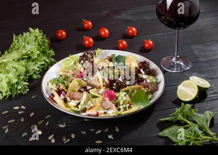 Salad with sliced meat, cheese, vegetables and salad mix on a white plate on a wooden table. Salad on a beautiful background with a glass of wine, lim Stock Photo