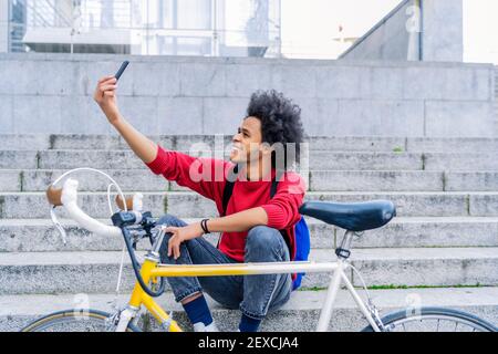 young man with afro hair sitting taking a selfie next to his old bicycle Stock Photo