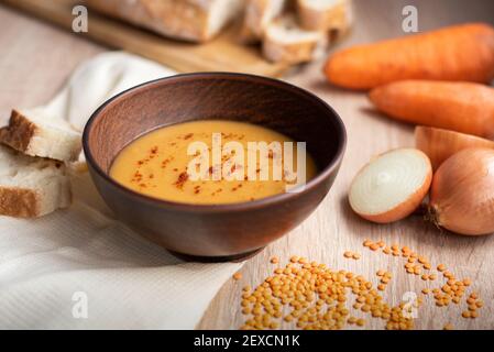 Hot delicious vegetarian red lentil cream soup with ingredients: carrot, onion, and bread on a wooden table, close up. Stock Photo
