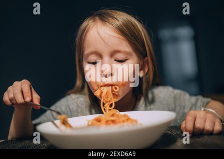 Hungry little girl appetite eating the bolognese pasta sucking it up to mouth in the low light kitchen table. Stock Photo