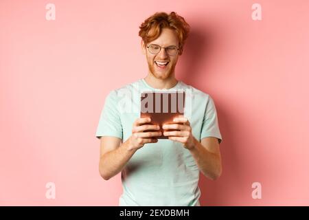 Happy guy with red hair and beard using digital tablet, reading screen and smiling, standing over pink background Stock Photo