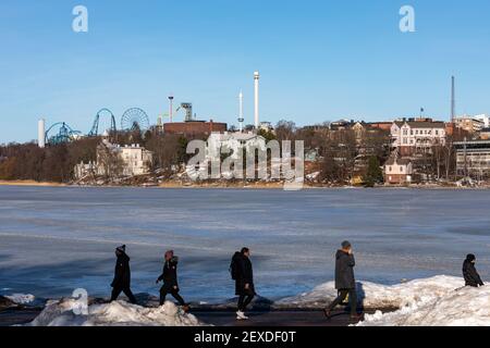 Frozen Töölönlahti Bay with passing people in front and Linnunlaulu villas and Linnanmäki amusement rides in the background in Helsinki, Finland Stock Photo