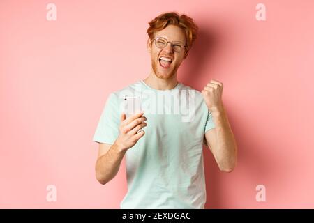Happy redhead guy in glasses and t-shirt winning online prize, shouting yes with joy and satisfaction, holding smartphone and making fist pump, pink Stock Photo