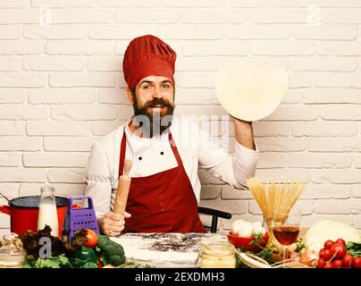 Chef makes pizza. Cooking process concept. Cook with excited face in uniform sits by table with vegetables holding rolling pin. Man with beard stretches pizza dough on white brick wall background. Stock Photo