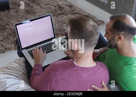 Multi ethnic gay male couple sitting on couch using laptop with copy space on screen at home Stock Photo