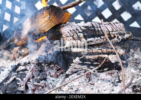 A fire in a caged metal fireplace burns outdoors, with white, glowing hot embers of the coal from burning wood. Smoke rises from the charred logs and Stock Photo