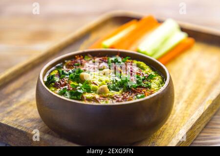 Homemade chickpea green hummus with herbs in round bamboo bowl, celery, carrot sticks on wood Stock Photo