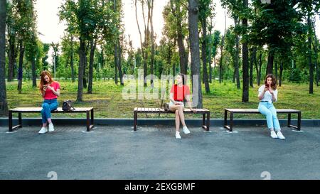 Social distancing, new normal concept. Three woman girlfriends with cell phone sitting on bench in park in social distancing. Social distance to avoid Stock Photo