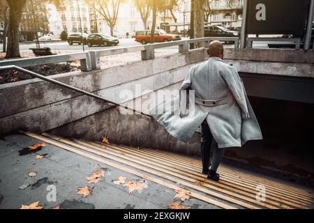 View from behind of a bald black guy in an elegant coat fluttering in the wind while he rapidly descending the stairs to an underground pass or a metr Stock Photo