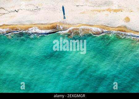 Aerial top view of single human figure casting shadow on sandy beach, standing by sea with beautiful azure tropical sea water and waves, copy space for your text. Stock Photo