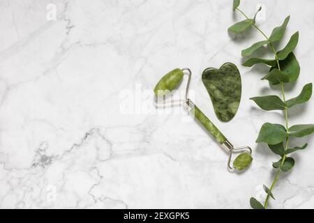 Green jade roller and gua sha stone for facial massage and eucalyptus branch on marble background. Home beauty and selfcare accessories, face roller Stock Photo