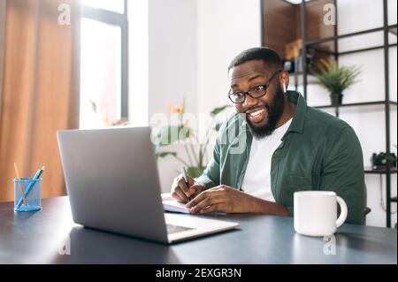 Distance learning at home. African American guy student or freelancer uses a laptop for learning, watches a webinar, makes notes in a notebook Stock Photo