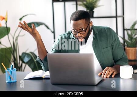 Dissatisfied confused multiracial black guy, male freelancer or manager, discouraged looks at the laptop, received unexpected message or news, sitting at workplace Stock Photo