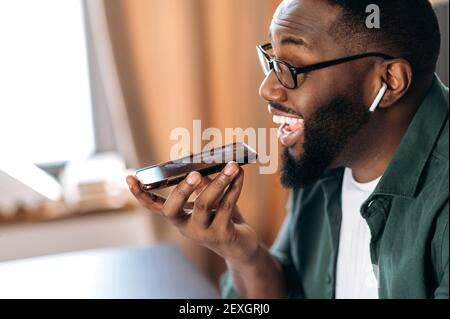 Happy smiling black guy, holding and using cell phone, speaking with friend or colleague speakerphone, sitting at home or office, record voice message Stock Photo