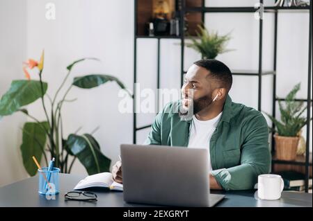 Confident african american young man sitting at work desk, using laptop for work, looking away and smiling, thinking about weekend or successful deal Stock Photo