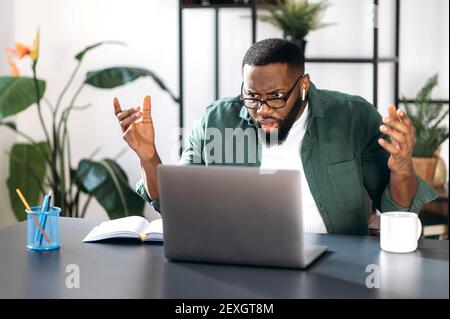 Dissatisfied confused multiracial black guy, male freelancer or manager, discouraged looks at the laptop, received unexpected message or news, sitting at workplace Stock Photo