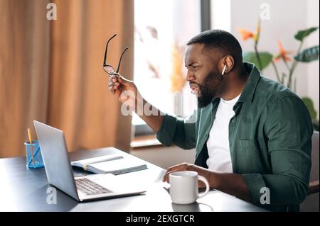 Dissatisfied confused multiracial african american male freelancer or manager, discouraged looks at the laptop, received bad message or news, sitting at workplace Stock Photo