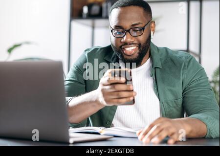 Chatting online. Joyful African American guy, sits at his desk, using a mobile phone while working, browsing the internet, answering an email, chatting with friend or family Stock Photo