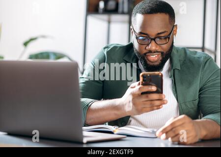 Busy confident African American guy, sits at his desk, using a mobile phone while working, browsing the internet, answering an email, looking for ideas for a project Stock Photo