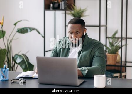 Concentrated serious young successful multiracial black ceo, programmer or freelancer working using laptop while sitting at his work desk Stock Photo
